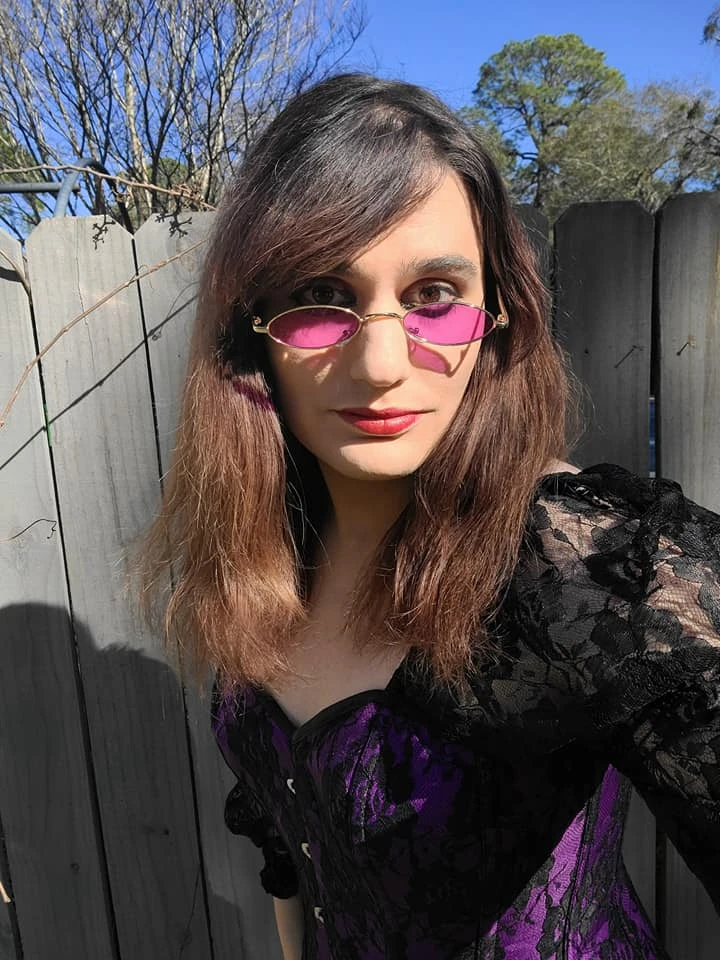 A person named Naia Ōkami is pictured outdoors in front of a wooden fence on a sunny day. Naia has shoulder-length brown hair and is wearing purple-tinted round sunglasses. Their makeup includes bold red lipstick, and they are dressed in a black and purple lace top. The background shows a clear blue sky and some trees.